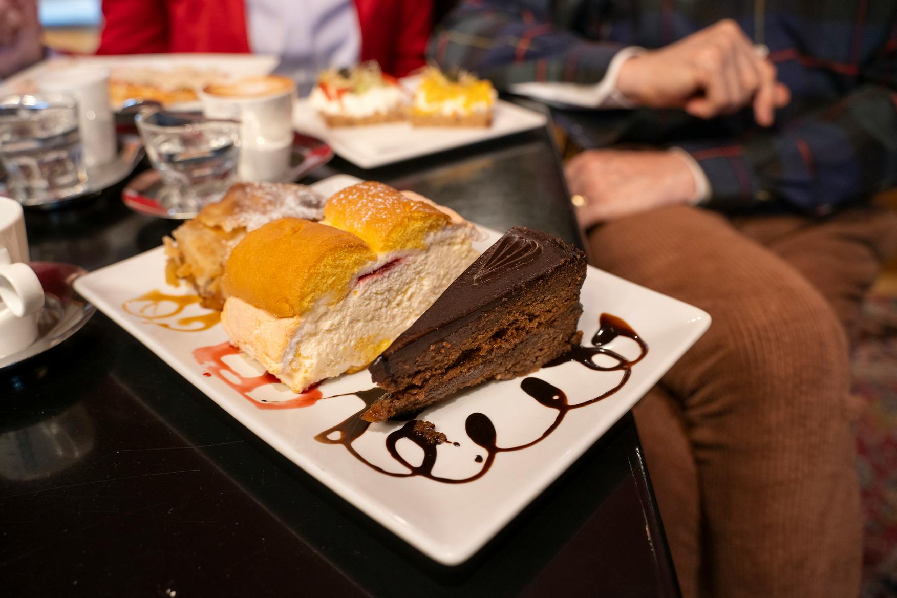 Um prato com bolo de chocolate e uma sobremesa em camadas, com pessoas sentadas nas proximidades e outros pratos visíveis em segundo plano.