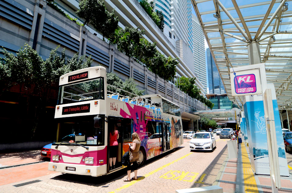 A colorful double-decker bus with "KL Hop" branding is at a city stop. People are boarding. Skyscrapers and a glass canopy are in the background.