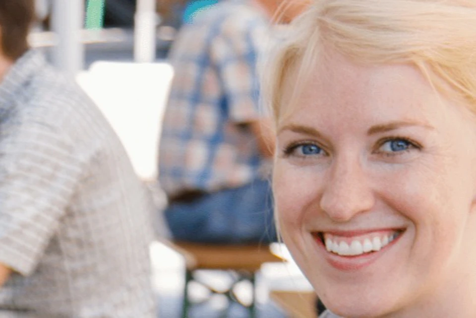 A smiling woman holding two large glasses of Löwenbräu beer at an outdoor seating area. People are seated and conversing in the background.