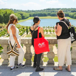 Three women stand on a terrace overlooking a formal garden with a pond and forest in the distance.