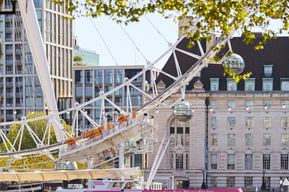 Urban scene with part of a Ferris wheel, glass capsules, modern buildings, and lush green trees. Boat on the river in the foreground.