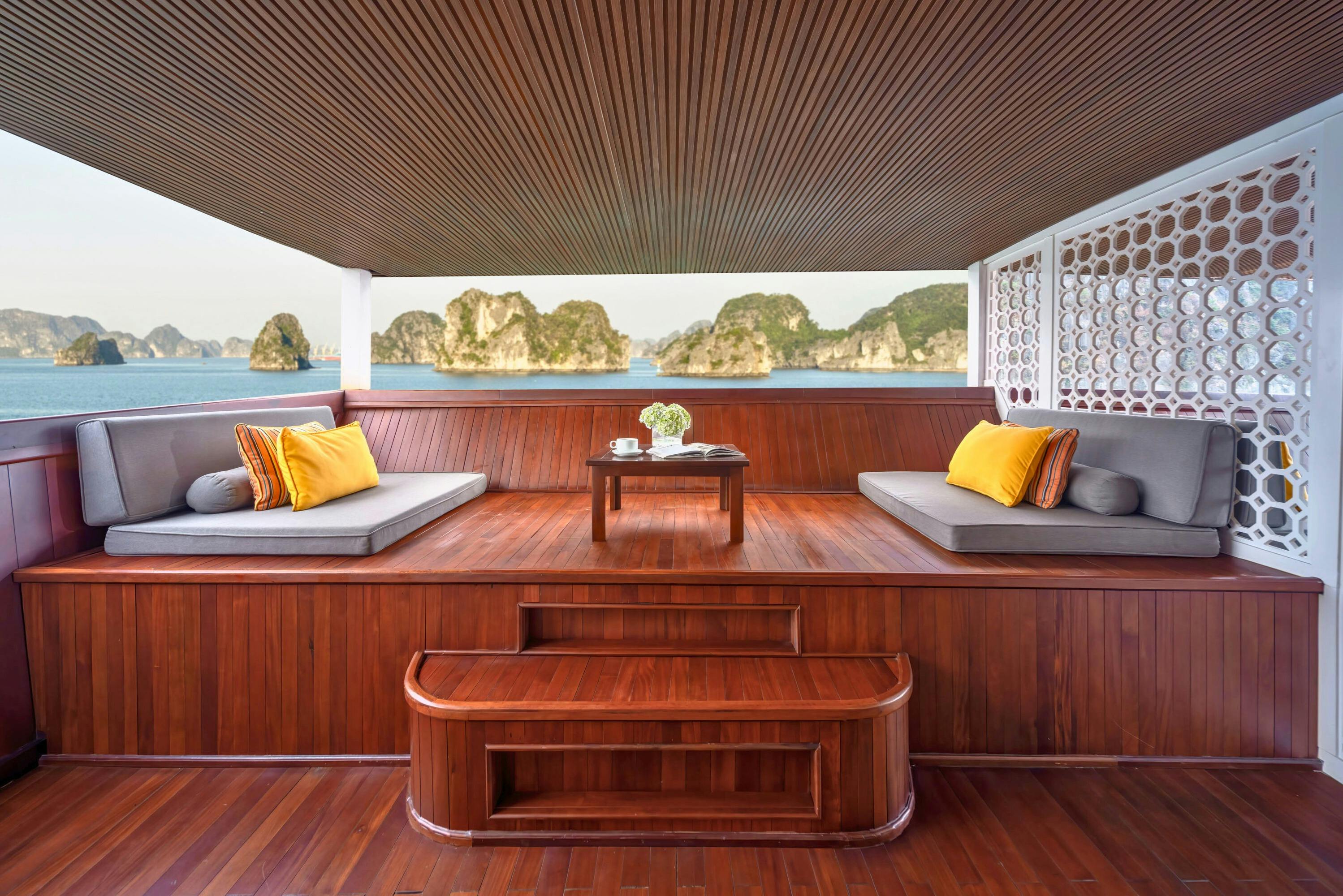 Wooden deck with cushioned seating, yellow pillows, and a small table with flowers, overlooking the ocean and rocky islands.