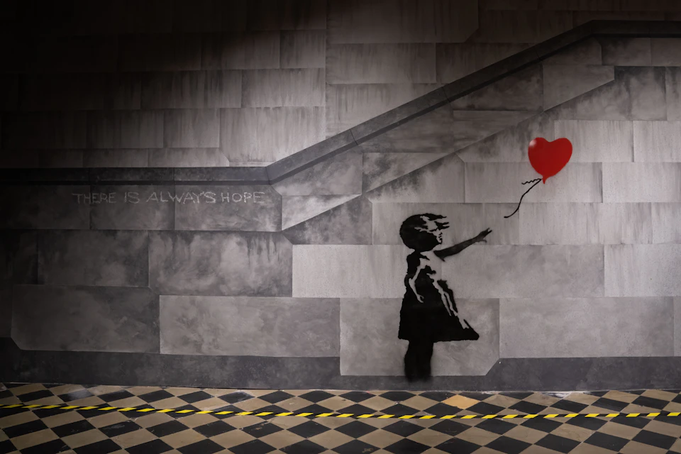 Stencil of a girl releasing a red heart-shaped balloon on a gray tiled wall with text "IS ALWAYS HOPE."