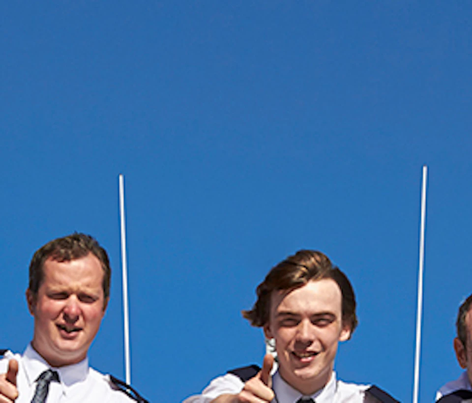 Five uniformed crew members on a vessel giving thumbs-up, standing behind a railing with a blue sky background.