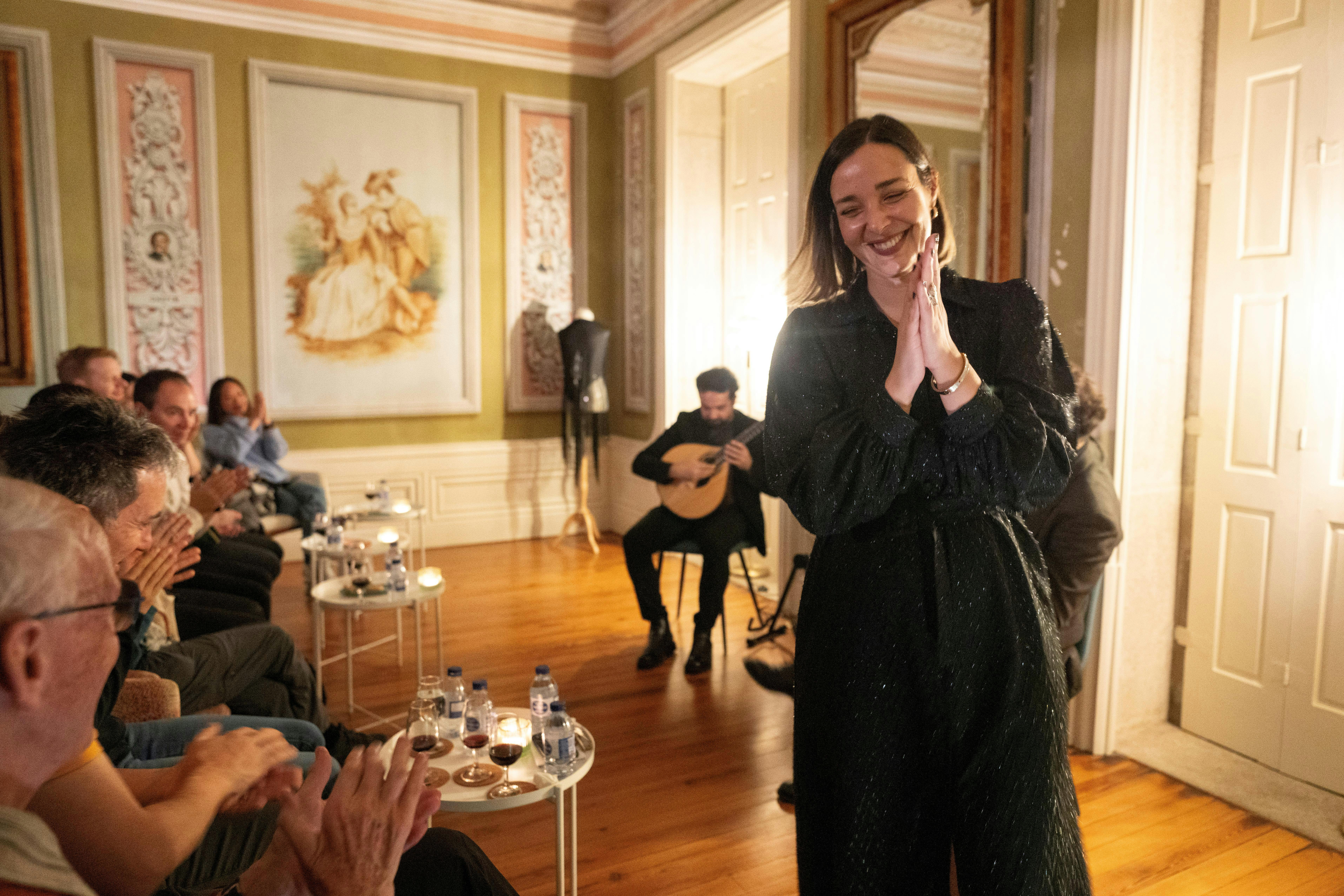 A woman in a black dress smiles with hands clasped, while a guitarist performs behind her in a warmly lit, elegant room.