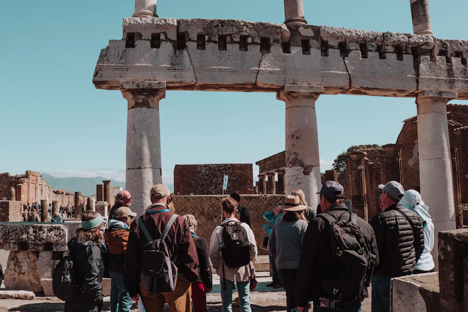 A group of tourists with backpacks and hats observe ancient stone ruins under a clear blue sky.