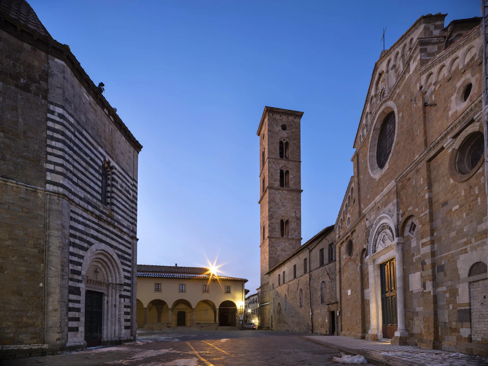 A quiet town square at dusk with historic stone buildings, a tall bell tower, and an ornate church facade under a clear sky.