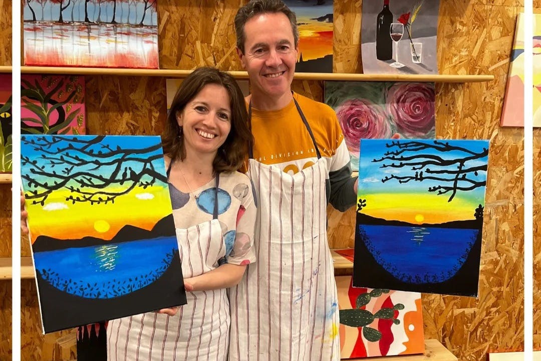 Δύο χαμογελαστοί άνθρωποι σε ποδιές κρατώντας πολύχρωμους πίνακες με ένα ηλιοβασίλεμα πάνω από το νερό, με φόντο διάφορα έργα τέχνης.