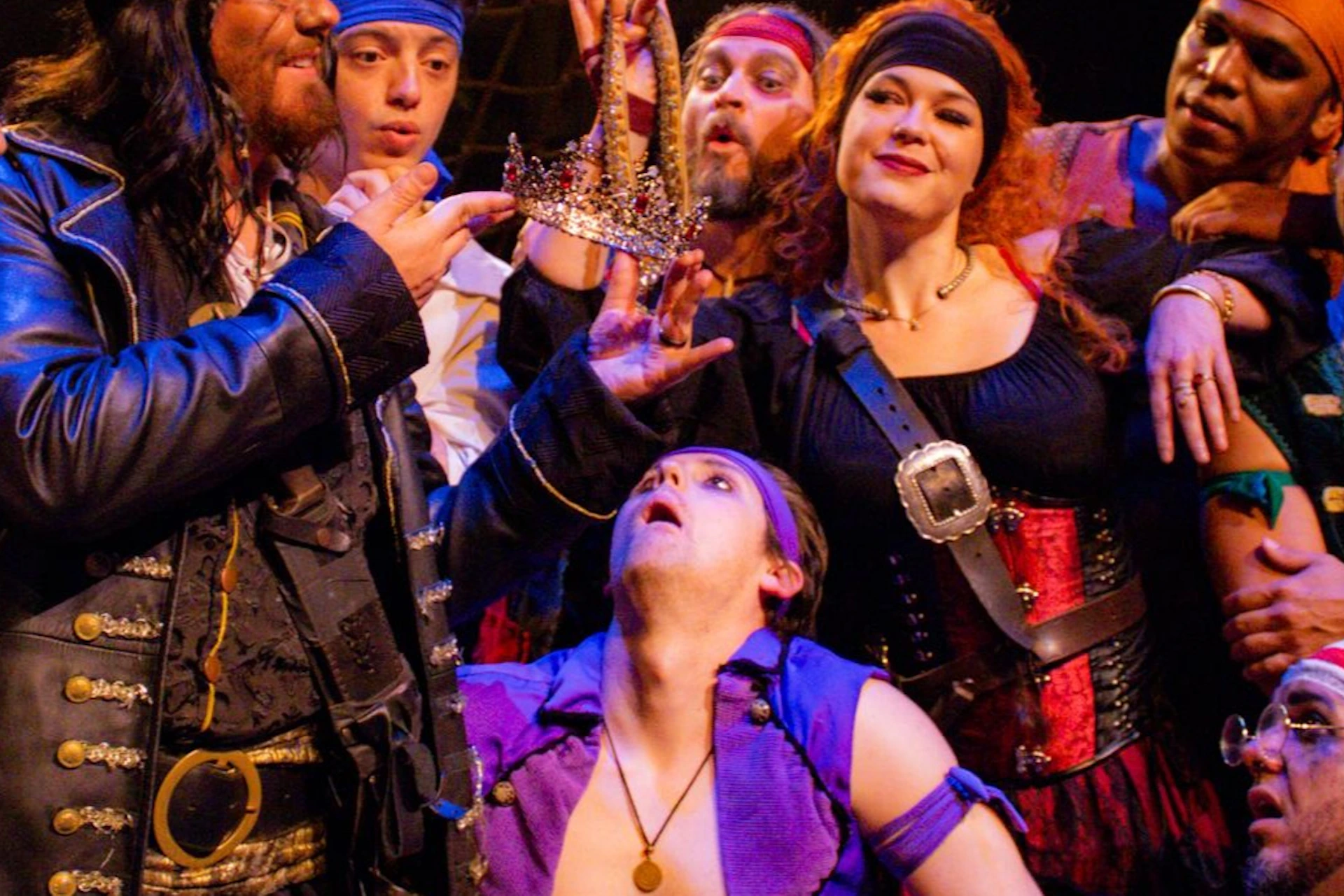 A group of people in pirate costumes gather around a man holding a crown, expressing excitement and curiosity.