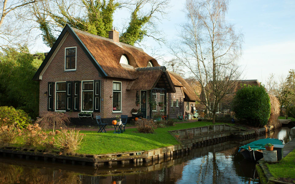 A brick house with a thatched roof next to a canal, surrounded by trees and grass, with outdoor furniture in the yard.
