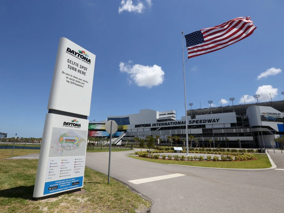 Daytona International Speedway entrance with a sign indicating a selfie spot, a large American flag, and a clear blue sky.