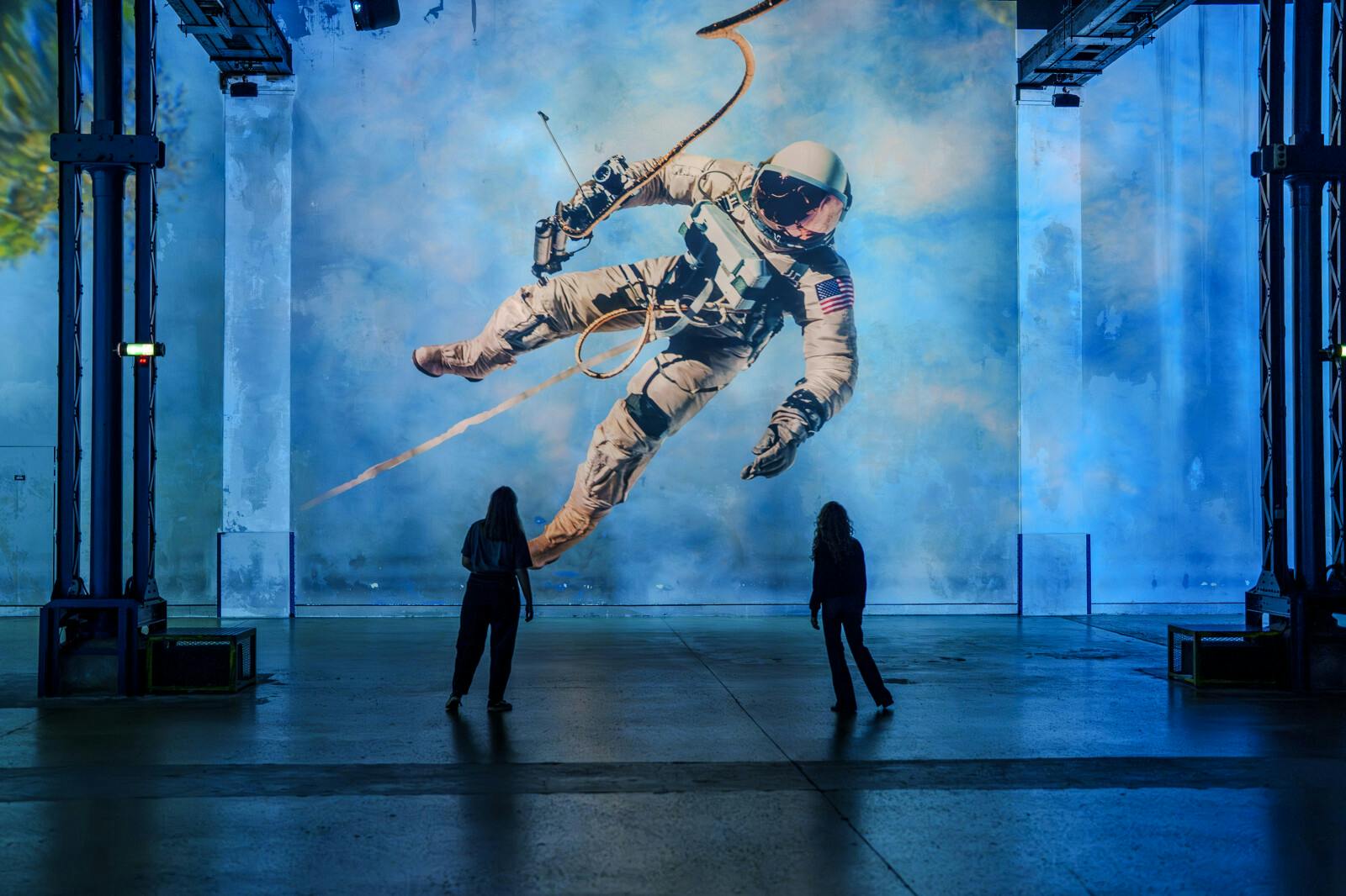 Δύο άνθρωποι στέκονται μπροστά από μια μεγάλη τοιχογραφία ενός αστροναύτη που επιπλέει στο διάστημα σε έναν μπλε τοίχο.