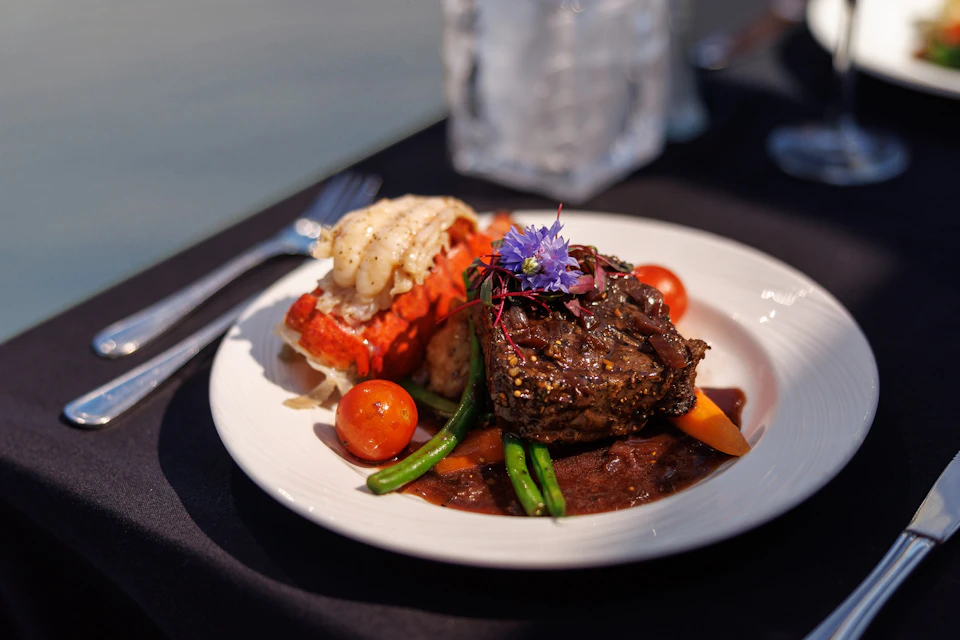 A gourmet plate featuring steak, lobster tail, cherry tomatoes, green beans, and garnishes, served on a white dish.