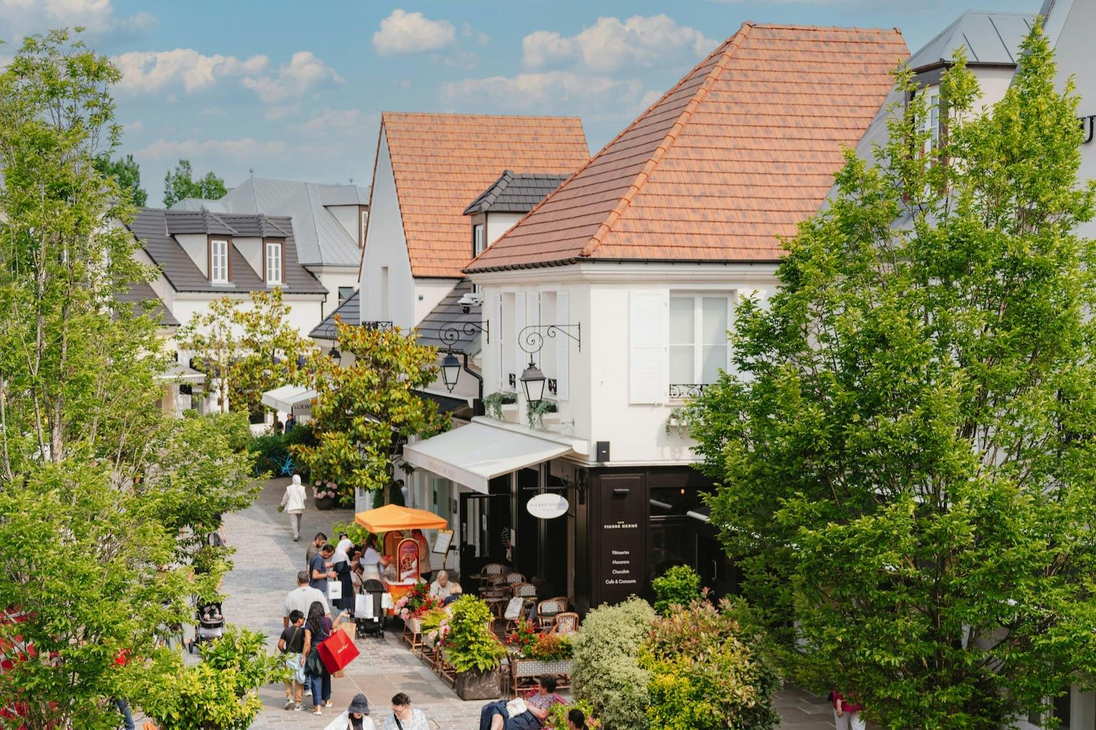 Ulica handlowa na świeżym powietrzu z ludźmi spacerującymi i siedzącymi w kawiarni, w otoczeniu zieleni i białych budynków z czerwonymi dachami.