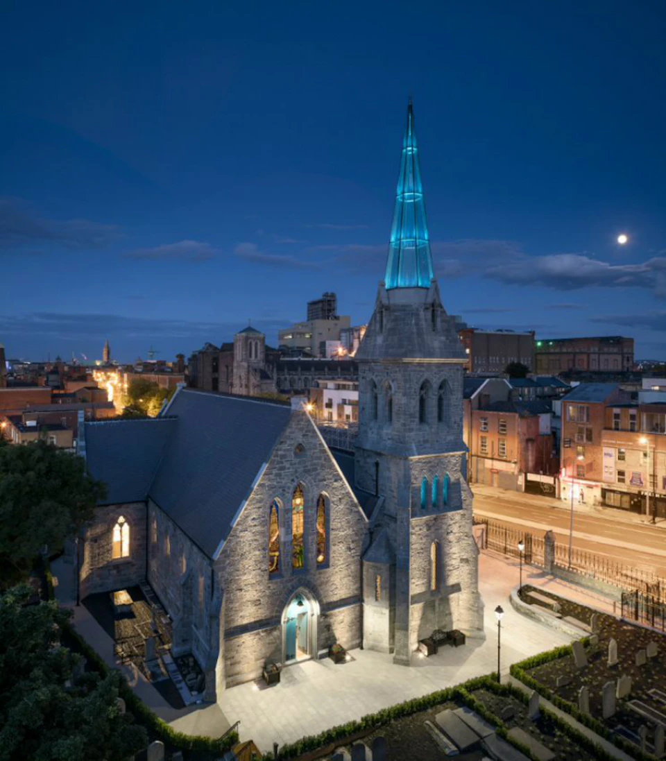 A stone church with a teal-lit steeple is illuminated at night, surrounded by urban buildings and streets.