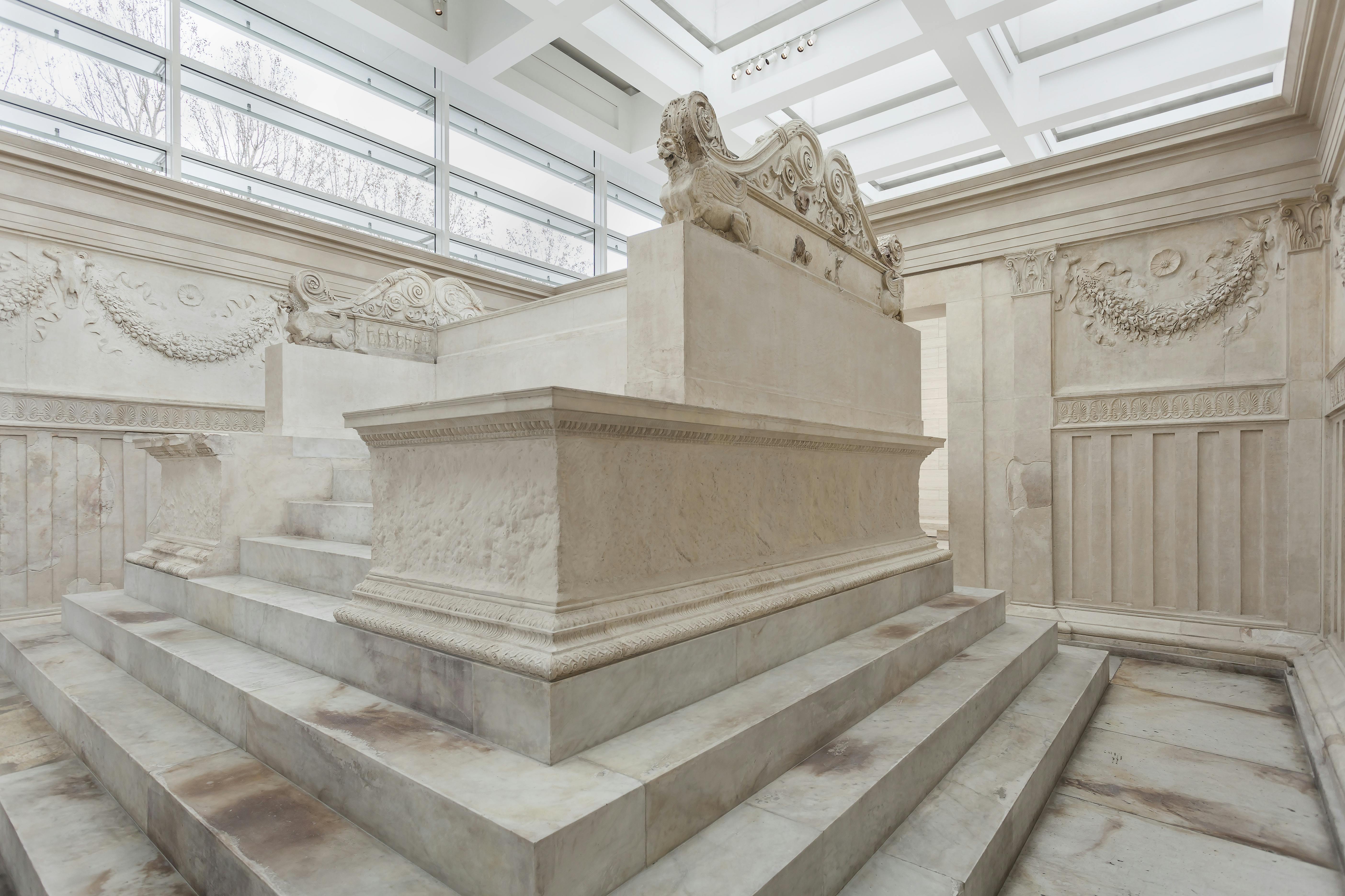 A large marble tomb with intricate carvings and sphinx sculptures, set on stepped platforms in a bright, modern museum.