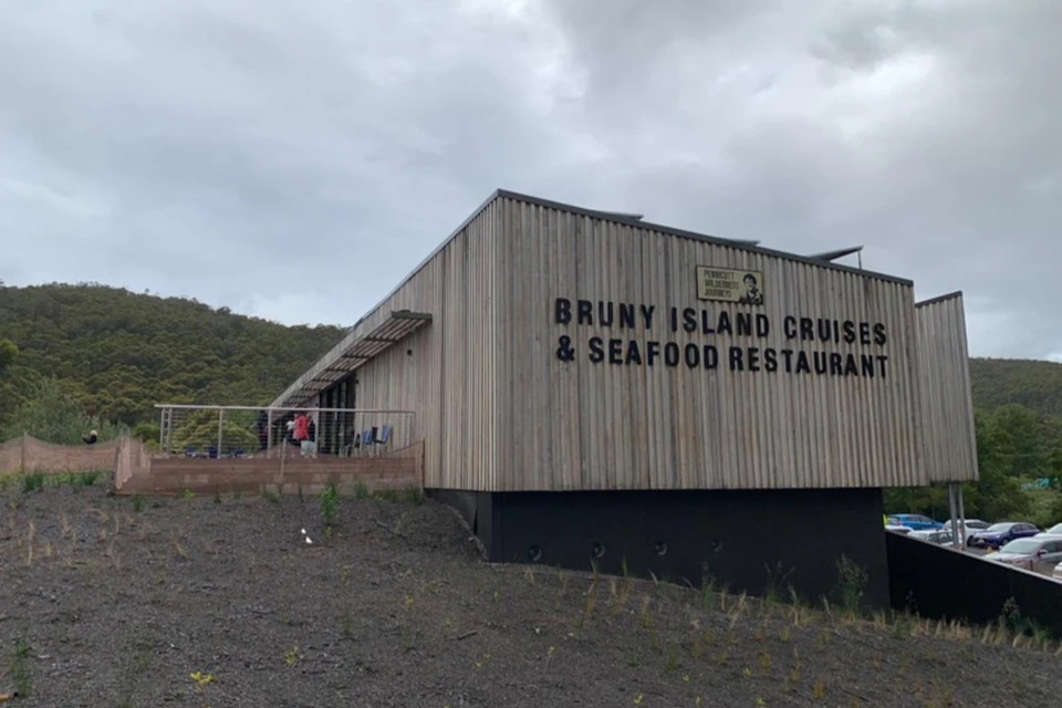 A wooden building with the sign 'Bruny Island Cruises & Seafood Restaurant,' set against a cloudy sky and greenery.
