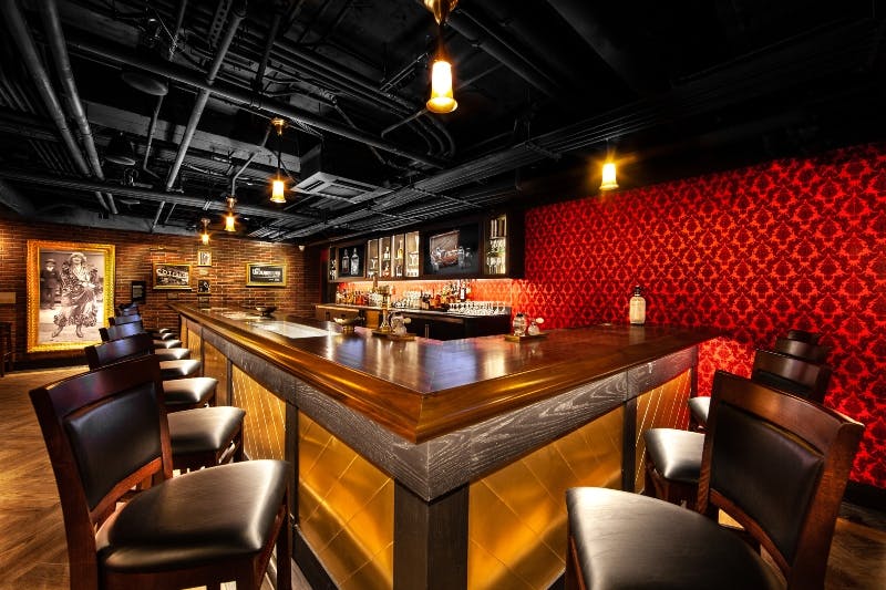 A stylish, dimly lit bar with a wooden counter, patterned red wallpaper, hanging lights, and rows of shelves with bottles in the background.
