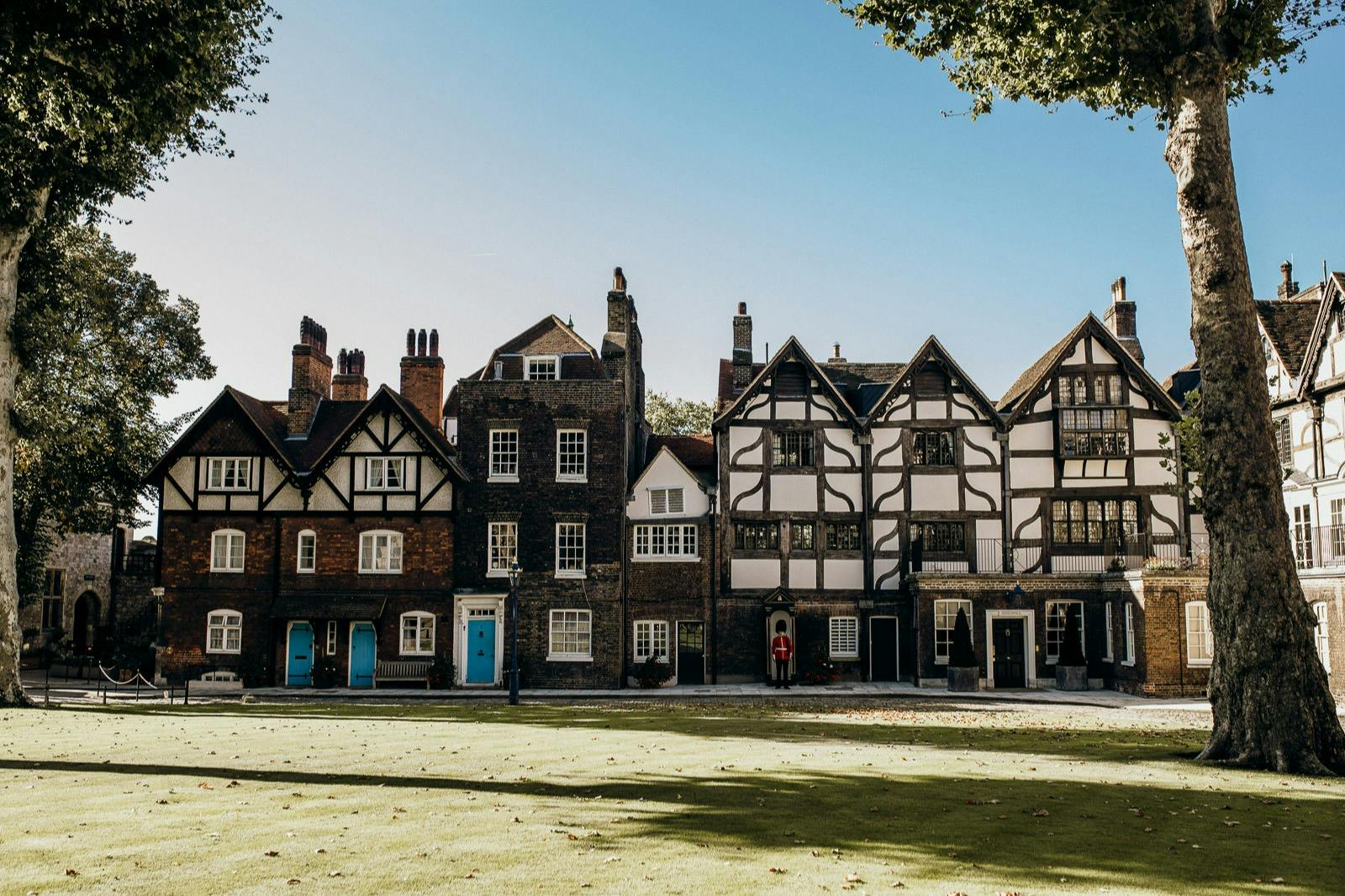Several connected, historic houses with various architectural styles and blue or white doors, set against a lawn and blue sky.