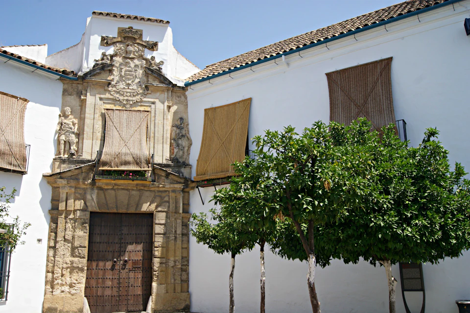 Historic stone building with wooden doors, ornate carvings, and a white facade. Trees and bamboo shades cover windows.