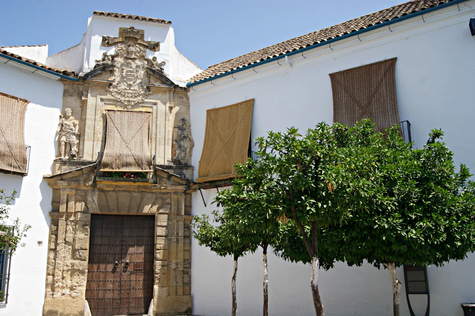 Historic stone building with wooden doors, ornate carvings, and a white facade. Trees and bamboo shades cover windows.