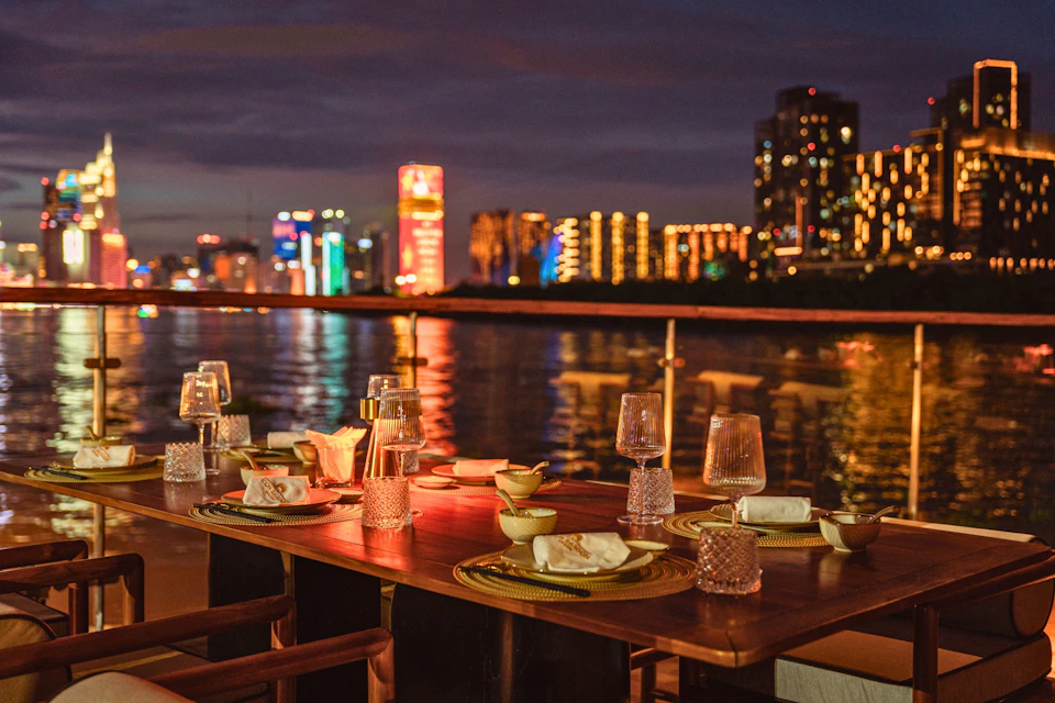 Outdoor dining table set with glasses, plates, and napkins, overlooking a cityscape with illuminated buildings at night.