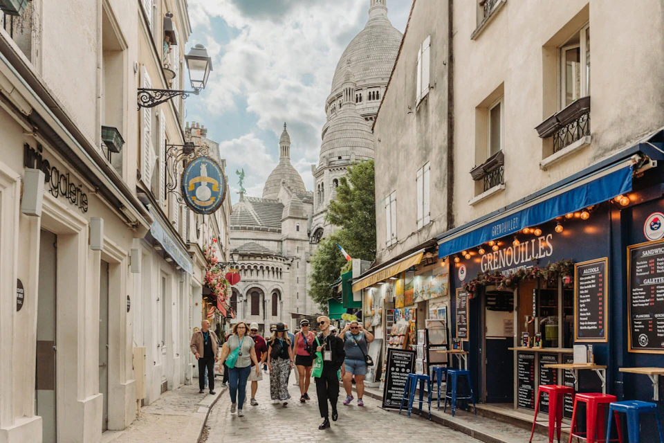 Tourists walking on a cobblestone street lined with shops and cafes, with Sacré-Cœur Basilica visible in the background.