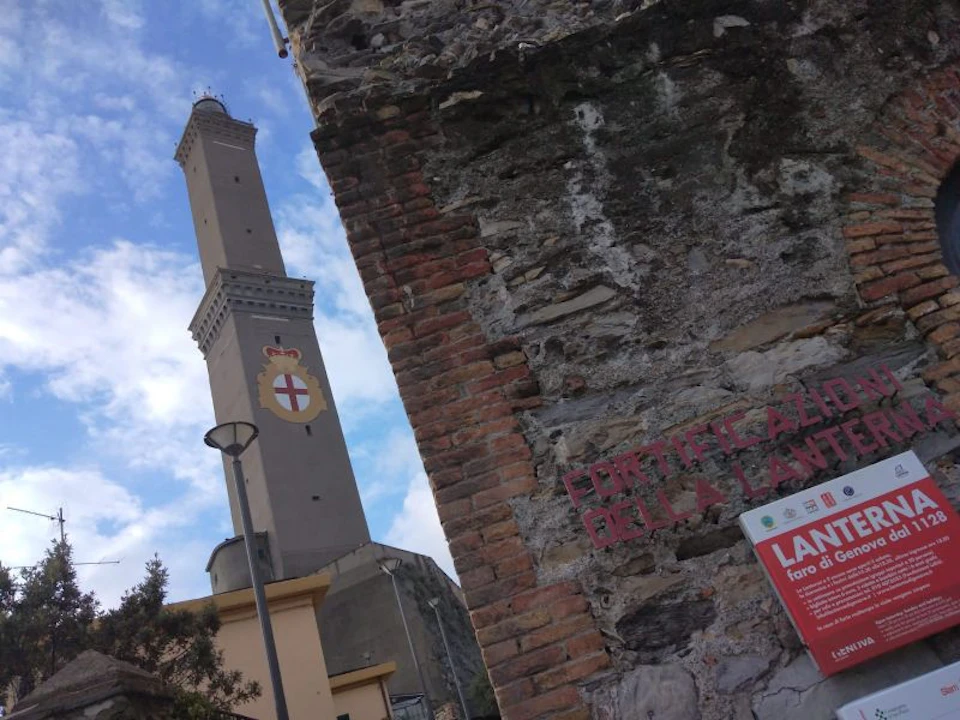 A tall lighthouse with a crest in the foreground, seen against a partly cloudy sky, next to old brick and stone walls.