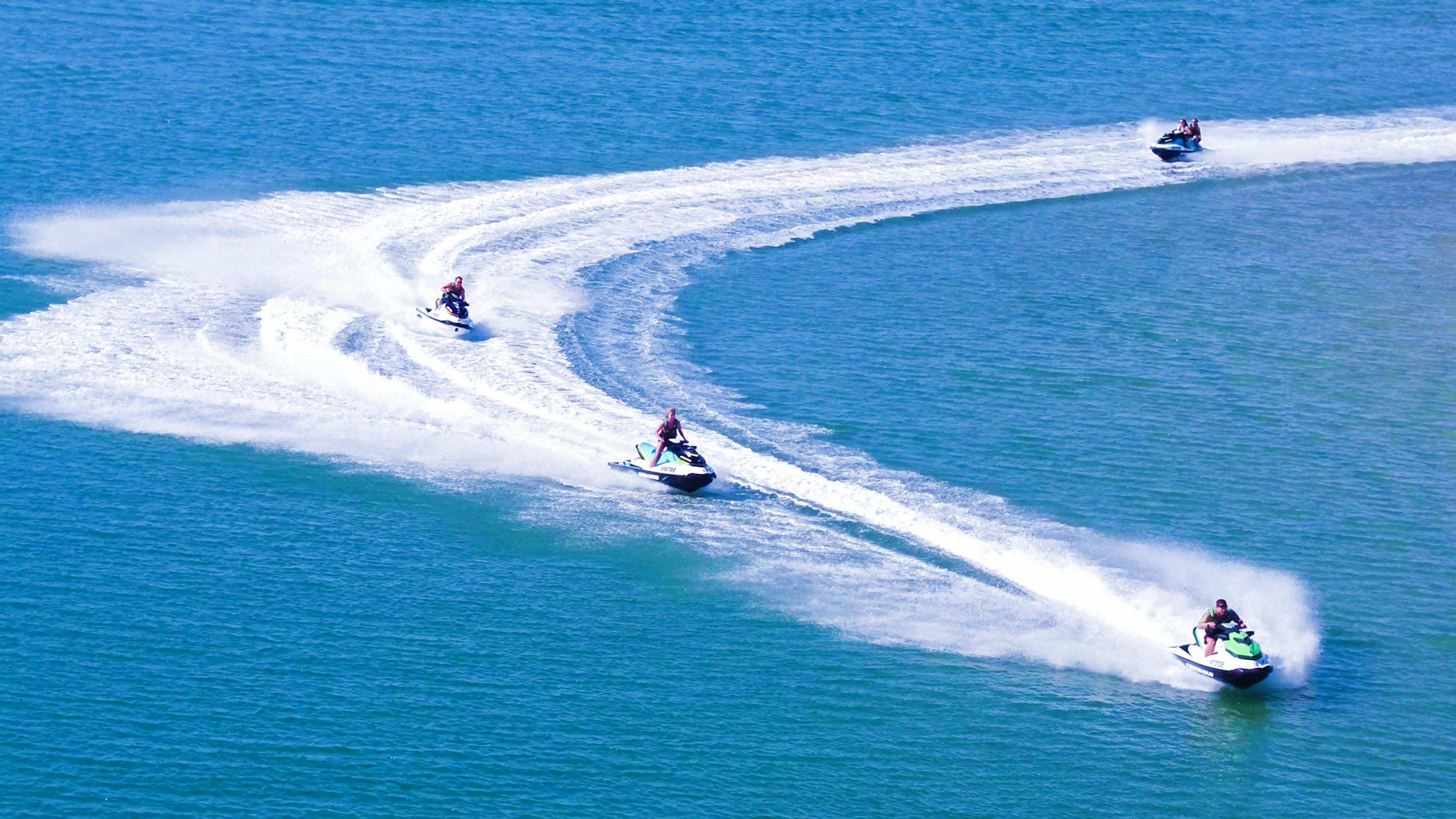 Two people riding jet skis create white trails on a large, calm body of blue water.