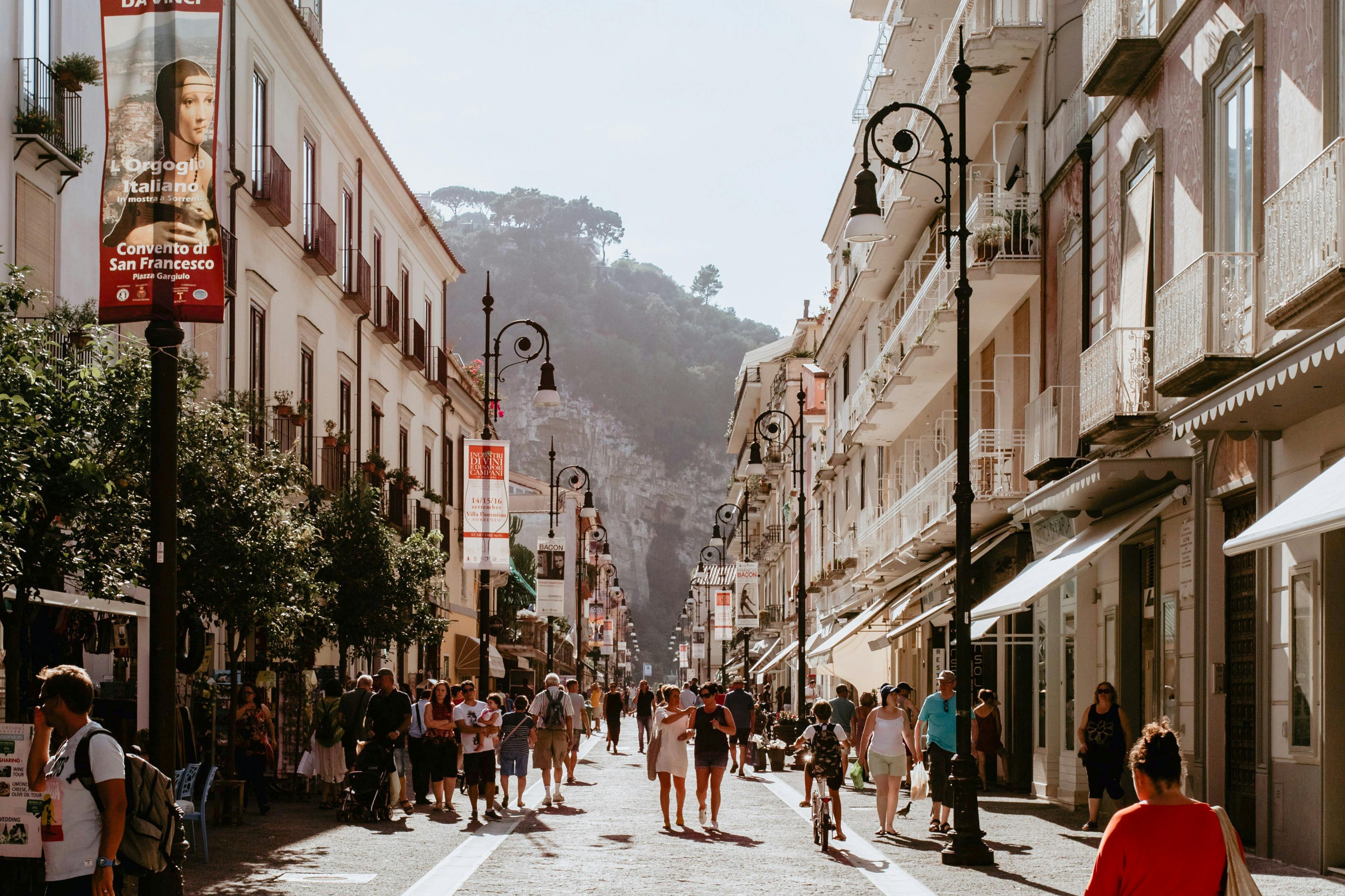 賑やかな歩行者専用道路には、建物や木々、街灯が並び、晴れた空の下を歩いたり、買い物をしたりする人々がいます。