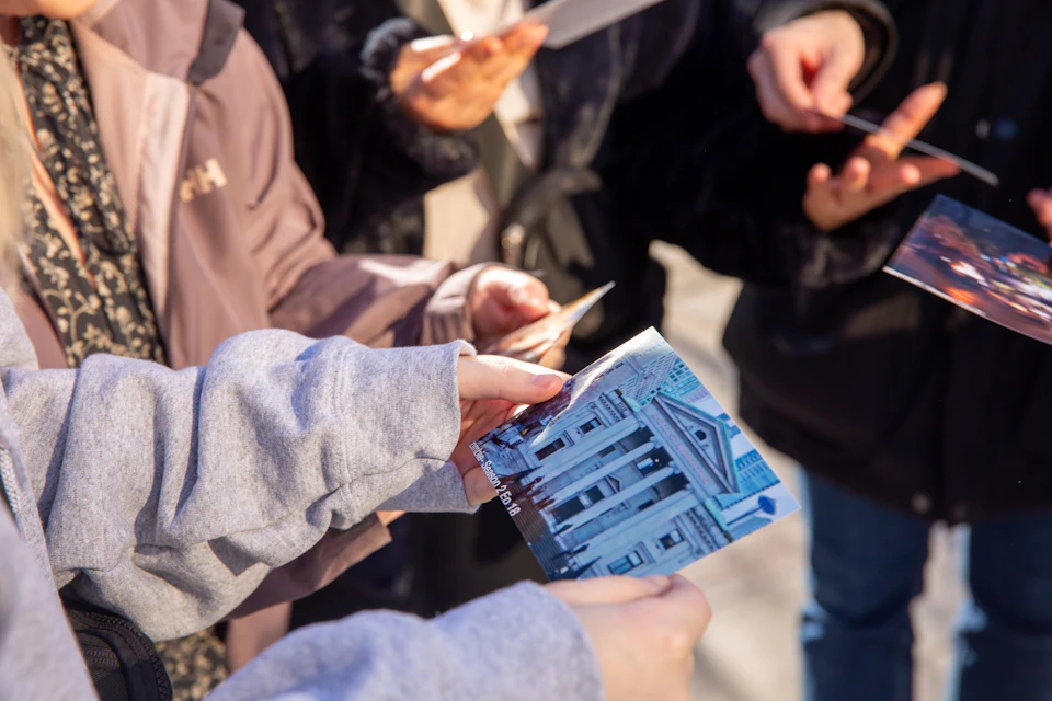 People holding printed photos and documents, one showing a building with columns and a triangular pediment.