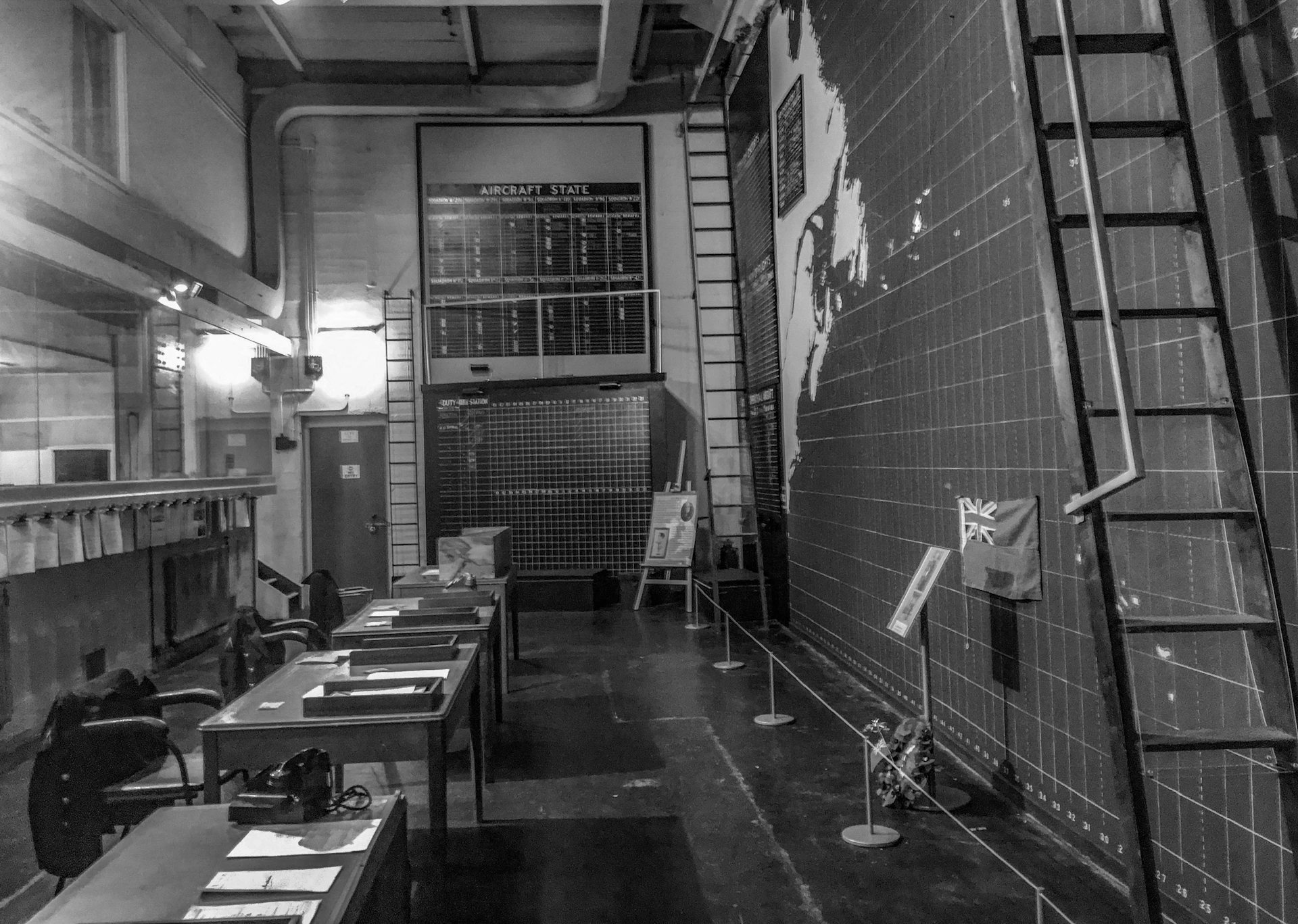 A black-and-white room with tables, ladders, large wall maps, and a board listing aircraft statuses. Flags and documents are on display.
