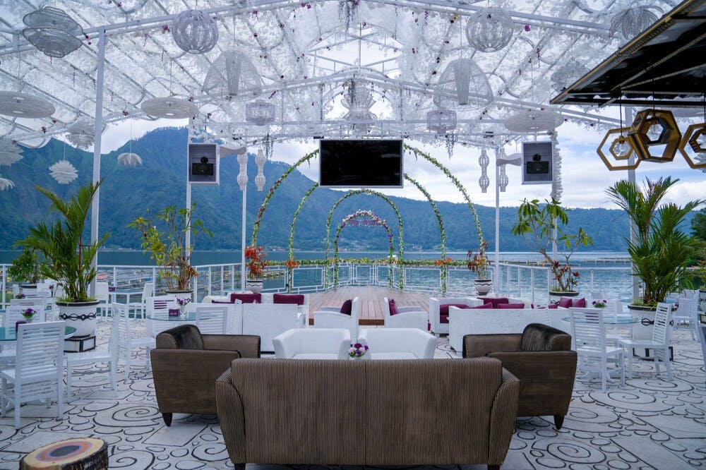 Outdoor lounge area with couches and chairs under a transparent canopy, overlooking a lake and mountains, with an archway and plants.