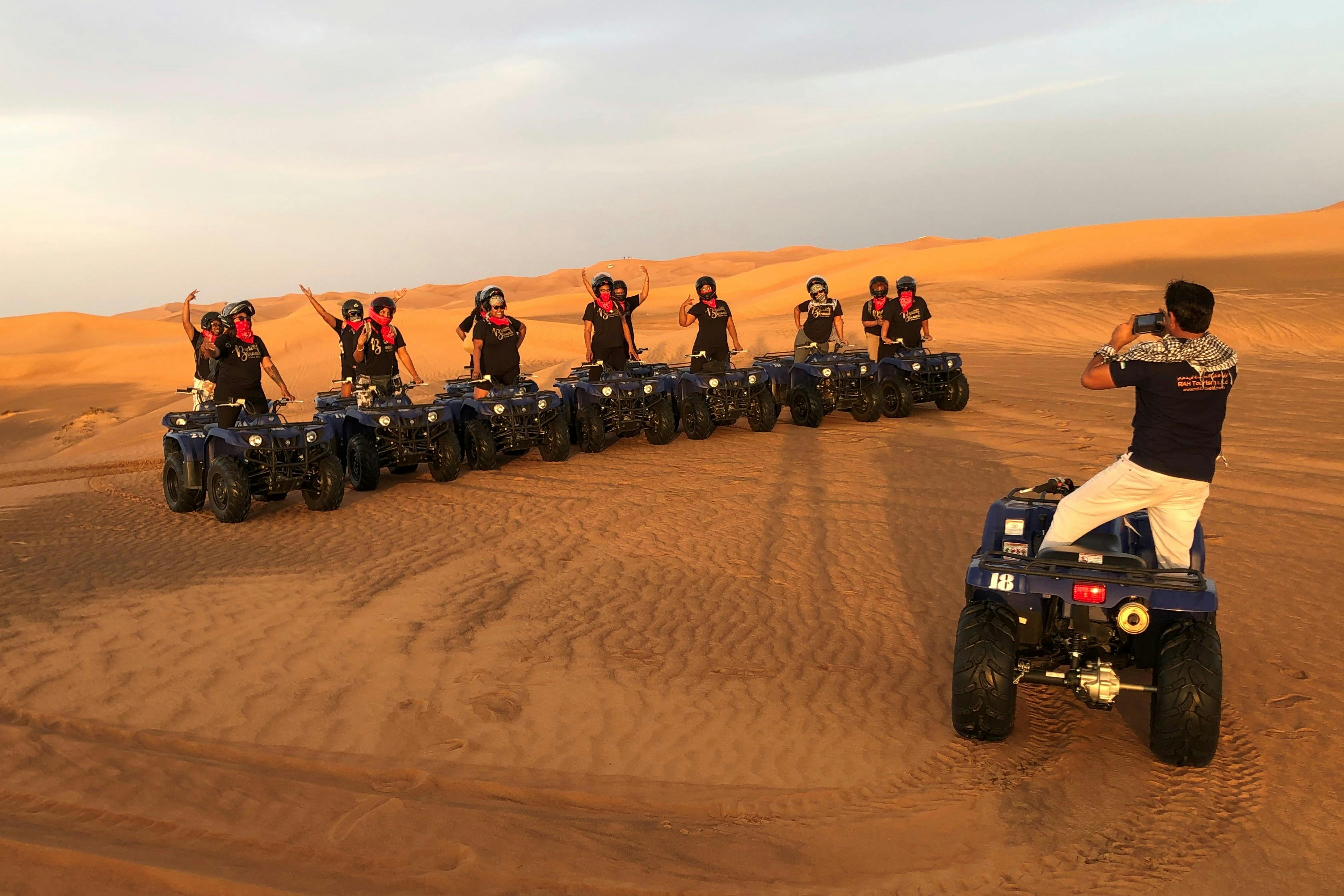 Lidé na čtyřkolkách stojí v řadě v poušti a zvedají ruce. V pozadí jsou vidět písečné duny.