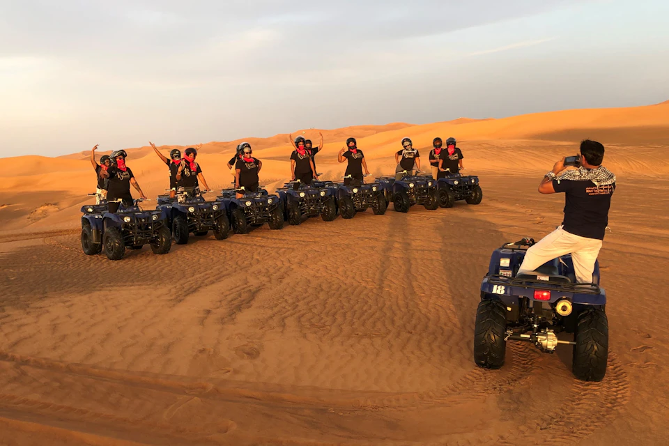 People on ATVs pose in a line in a desert, raising their arms. Sand dunes are visible in the background.