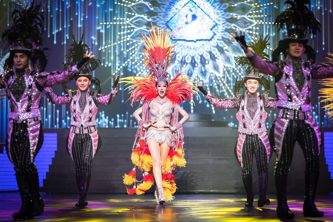 Performers in glittering costumes dance on stage; the central figure wears a feathery headdress and vibrant red and yellow plumes.