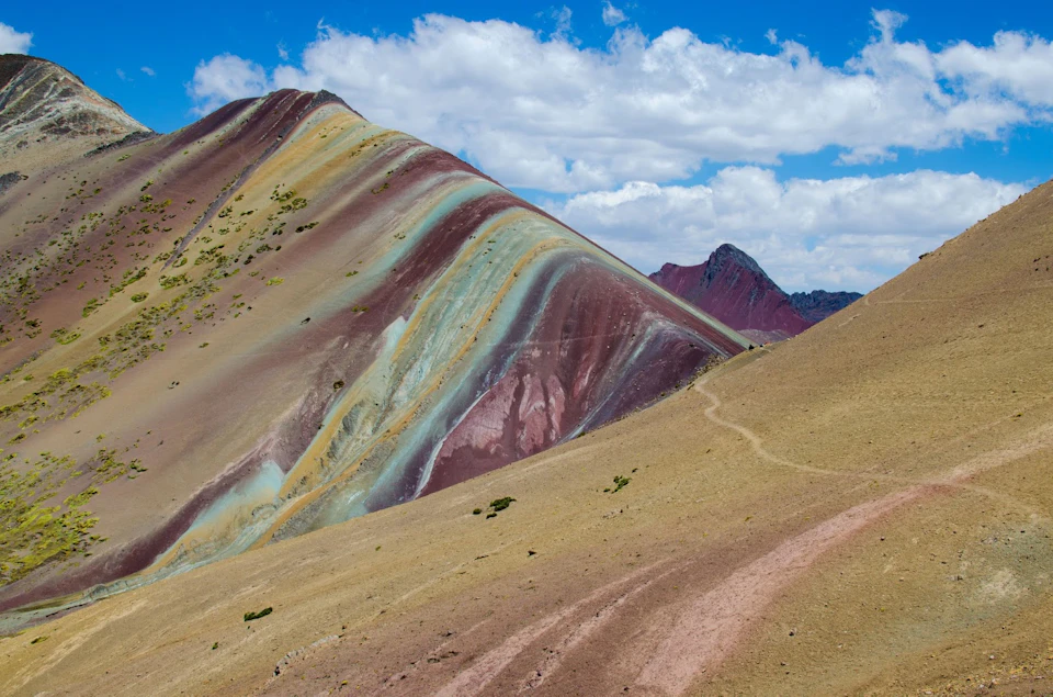Colorful mountain range with hues of red, yellow, and green beneath a partly cloudy sky. A narrow path winds through the landscape.