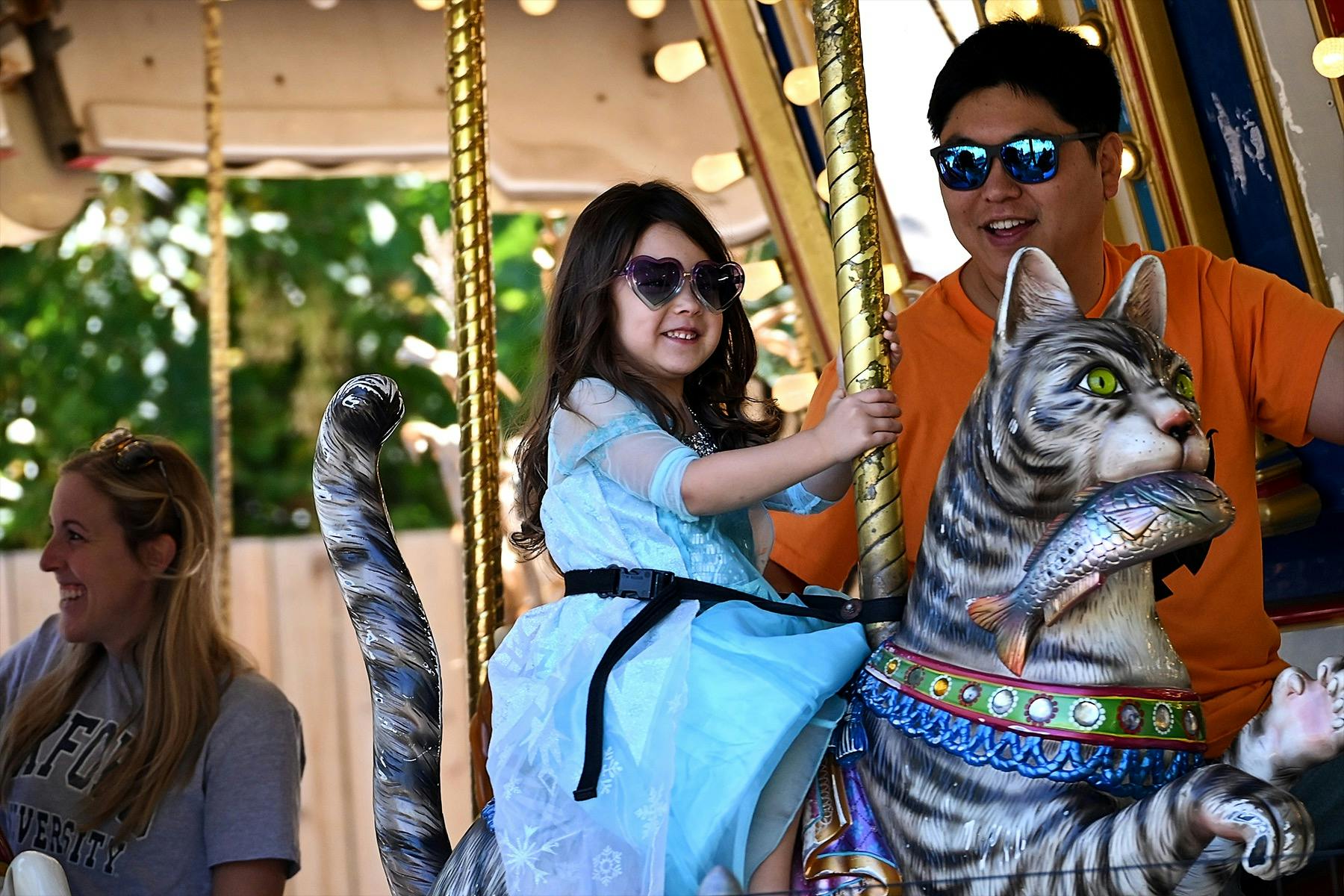 A young girl wearing sunglasses and a blue dress rides a carousel horse, accompanied by two adults, amid a sunny outdoor setting.