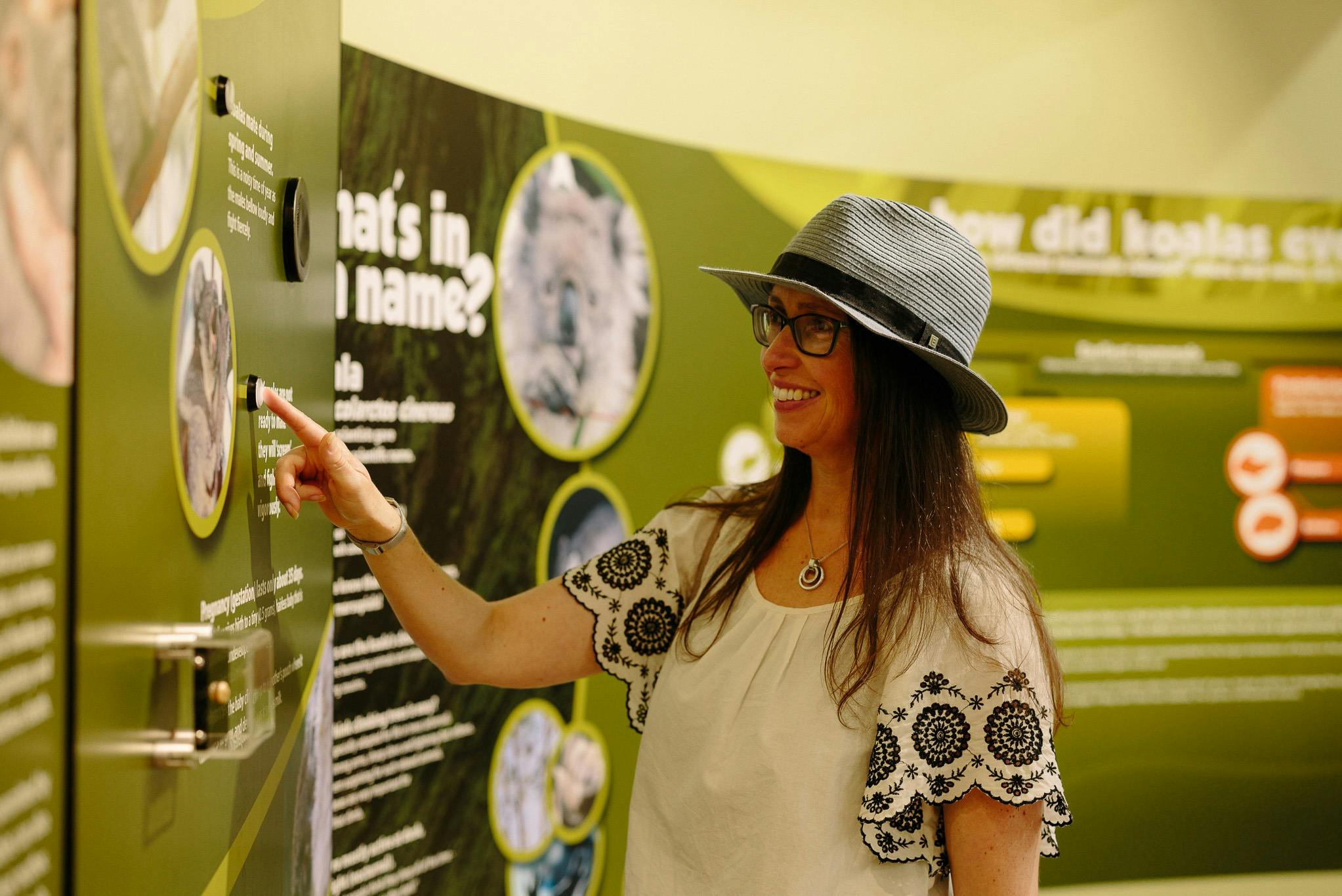 A woman wearing a hat and glasses smiles while pointing at a display in a museum or similar educational setting.