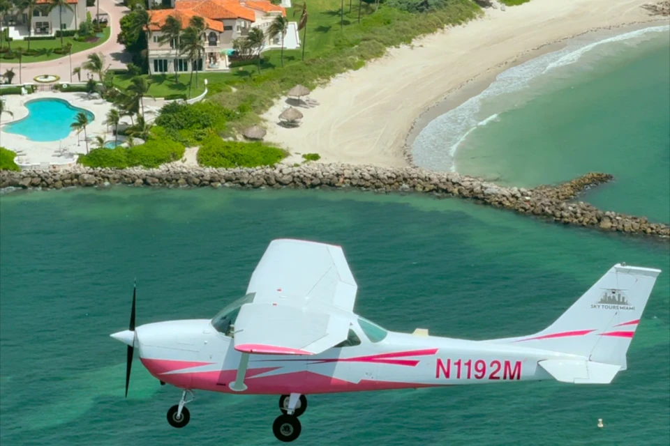 A white and pink propeller plane flies over turquoise water near a sandy beach with a house and palm trees in the background.