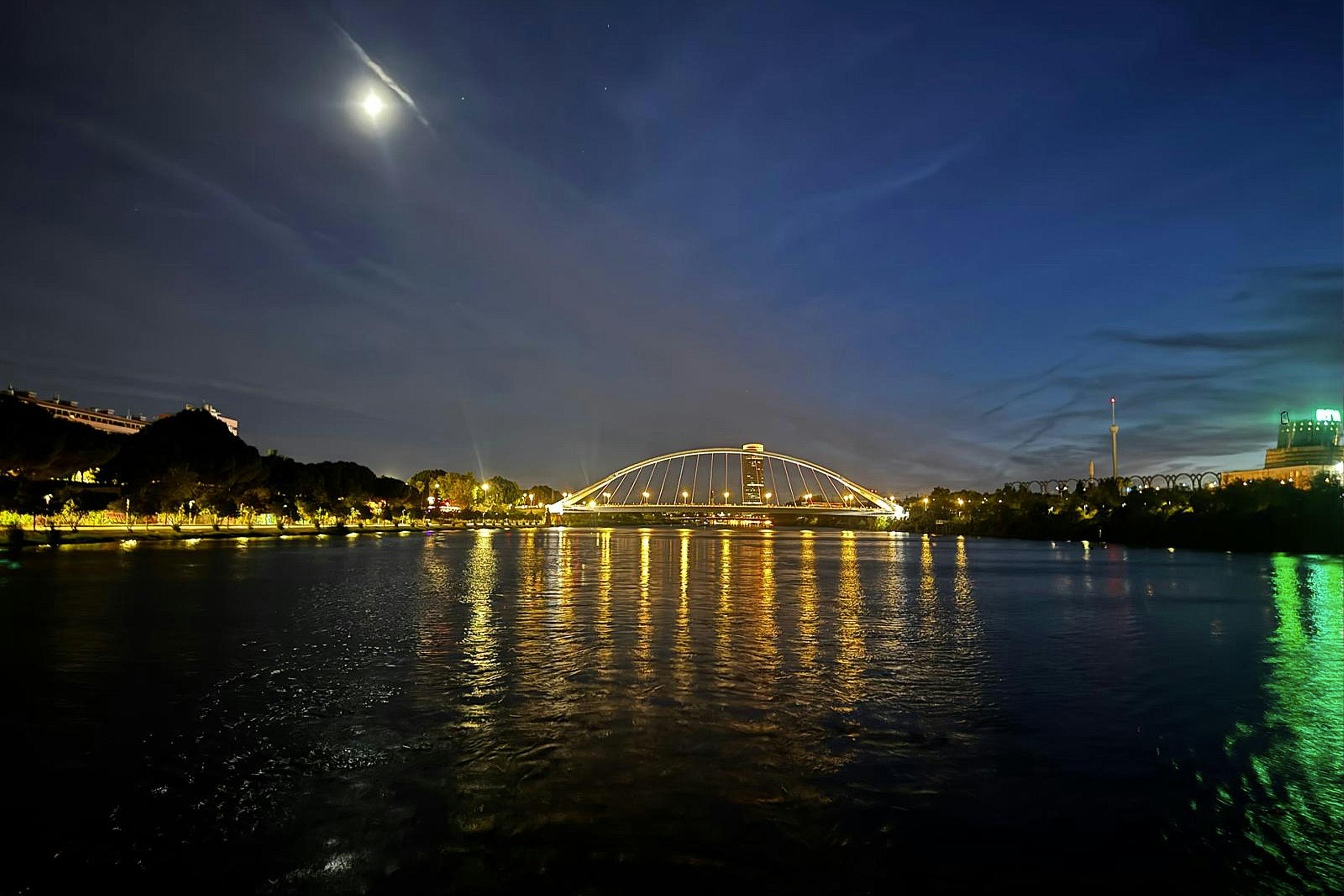Podświetlany most nad rzeką w nocy z odbiciami w wodzie i jasnym księżycem na częściowo zachmurzonym niebie.