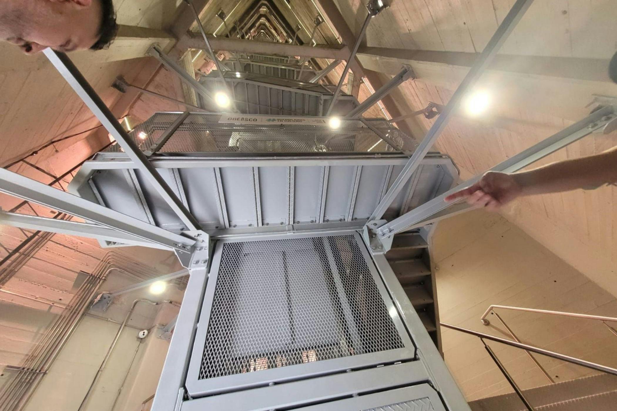 Looking up inside a metallic structure with multiple levels, beams, and bright lights. A hand points towards the structure.