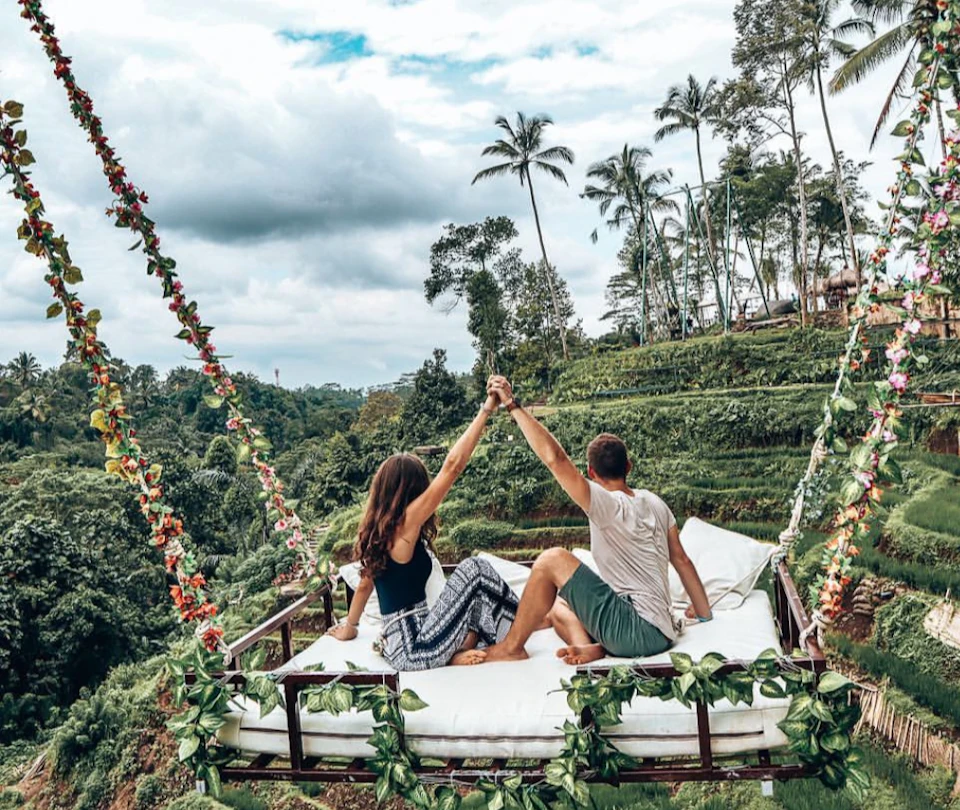A couple sits on a decorated swing adorned with flowers, overlooking a lush, green terraced landscape under a cloudy sky.