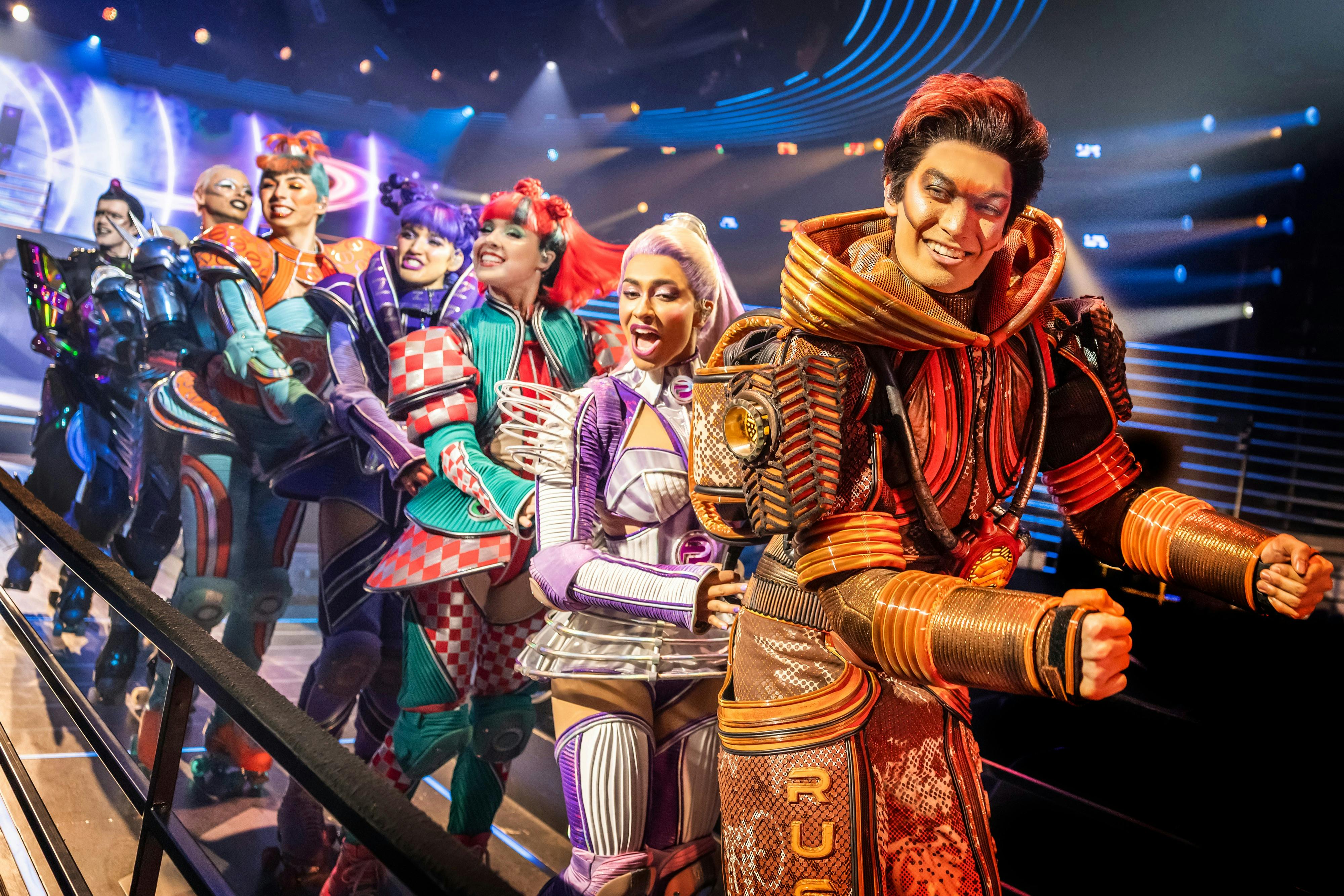 穿着精致、未来主义服装的人们在灯光明亮的舞台上表演，聚光灯色彩缤纷。