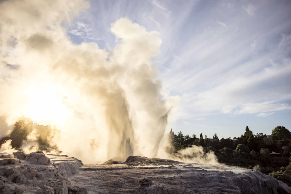 A geyser erupting with steam and water under a cloudy sky, surrounded by rocky terrain and distant trees.