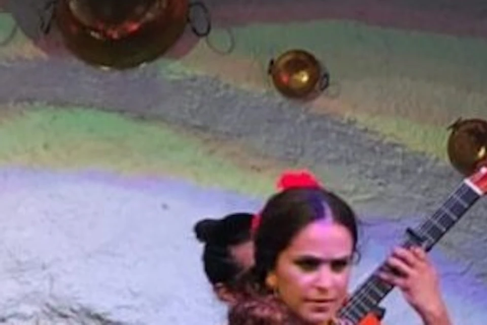 A flamenco dancer performs on stage with two women clapping on either side. The background features arched walls and decorative plates.