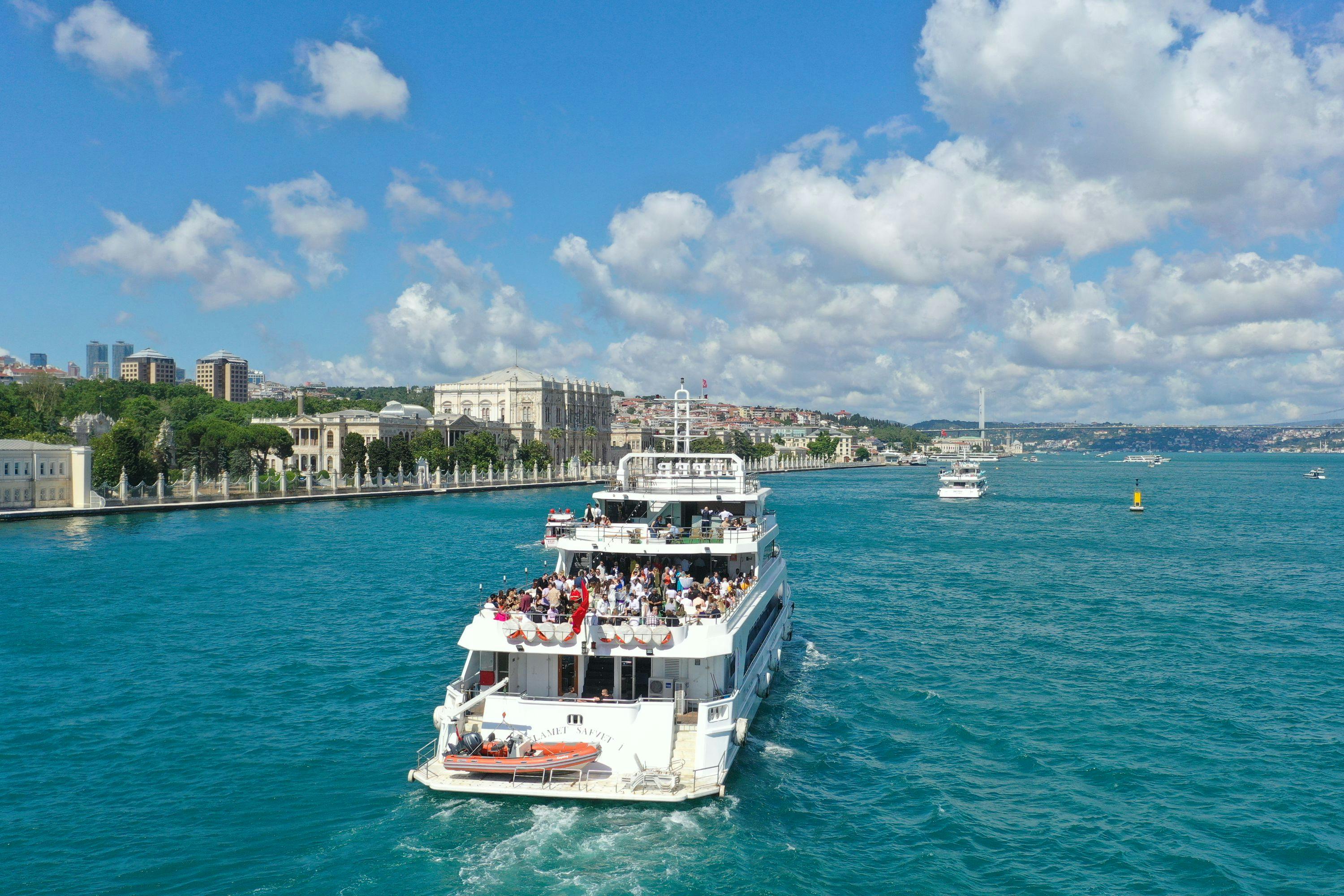A white passenger boat full of people travels on a blue waterway near a city with historic buildings and a clear sky.