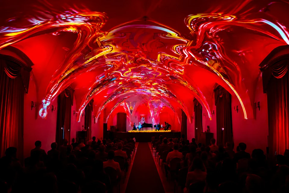 A concert in a hall with a vivid red and orange illuminated, ornate ceiling, and an audience seated facing a stage.