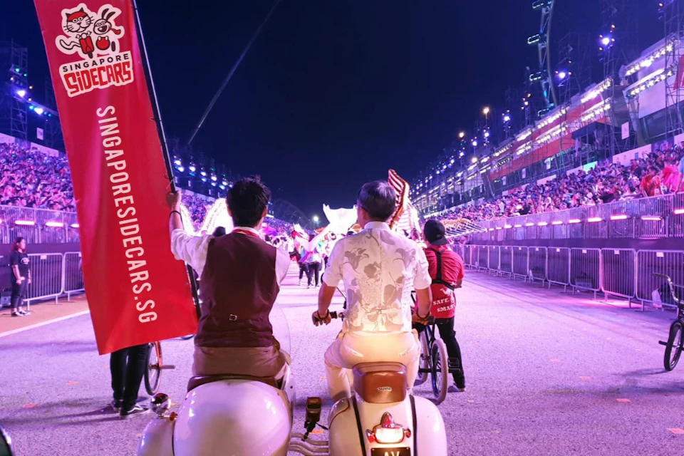 Two people riding a vintage sidecar and bicycle on a lit-up street at night, holding a red banner. Crowds are visible in the background.