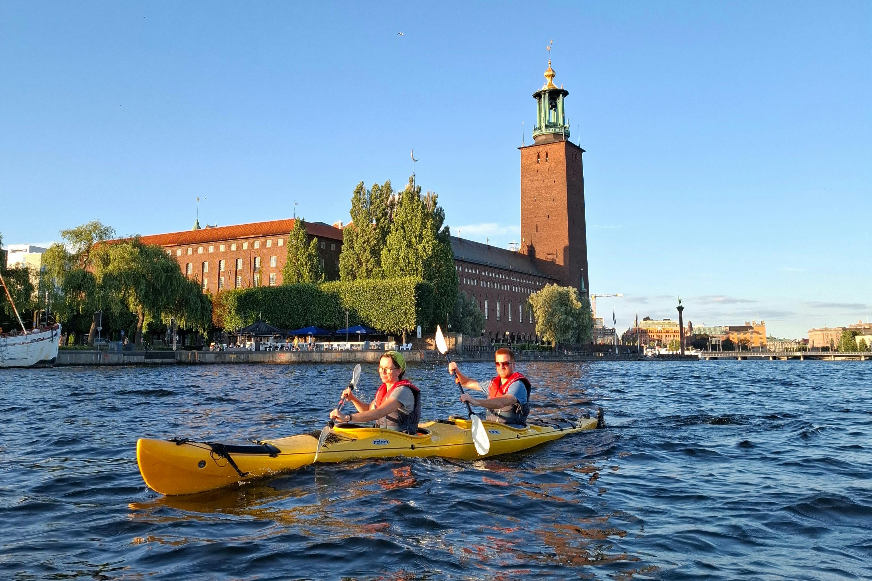 Deux personnes dans un kayak jaune pagaient sur une large rivière, avec un grand bâtiment en briques et une tour visibles à l'arrière-plan.